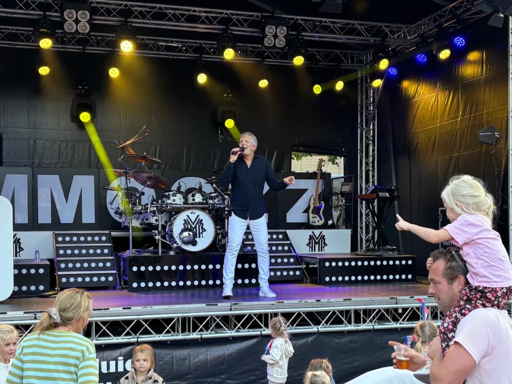 Feestzanger Marcel zingt op een podium met gele spotlights en een drumstel, met kinderen en publiek op de voorgrond in Hardenberg